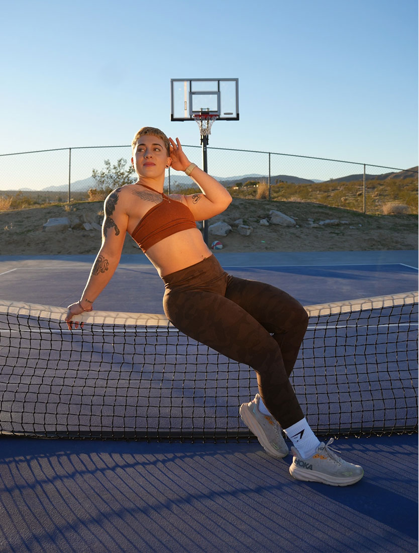 The coach sits against a court net outdoors with a basketball hoop behind her.